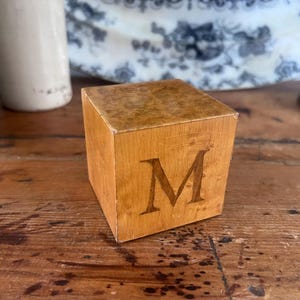 May include: A wooden cube block featuring a natural light brown wood grain finish and a dark brown letter M engraved on the front face. The block has a smooth, matte texture and sharp, defined edges, showcasing the organic patterns of the wood.