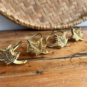 May include: Four gold-coloured leaf-shaped napkin rings. The rings are made of metal and have a detailed leaf design. The napkin rings are on a wooden surface with a woven basket in the background.