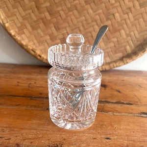 May include: A clear, cut-glass sugar bowl with a lid and a small spoon. The bowl has a diamond pattern and a round base. The lid has a knob on top. The spoon is silver. The bowl is sitting on a wooden surface.