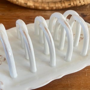 May include: A white ceramic toast rack with six arched slots, set on a scalloped dish. The rack has subtle brown accents and a floral design on the base, perfect for serving toast.