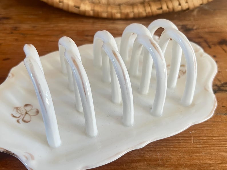 May include: A white ceramic toast rack with six arched slots, set on a scalloped dish. The rack has subtle brown accents and a floral design on the base, perfect for serving toast.