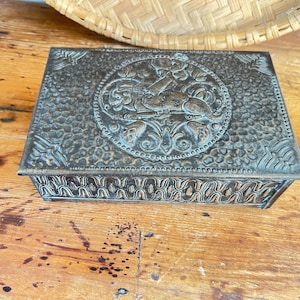 May include: A rectangular, antique-style metal box with a detailed, embossed design. The lid features a central circular emblem with a figure and floral motifs. The box has a textured surface and decorative trim, suggesting it's for jewelry or keepsakes.