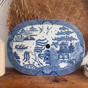 May include: An oval ceramic platter insert featuring a blue and white Willow pattern with a central drainage hole and numerous smaller perforations across the surface, depicting traditional pagodas, willow trees, a bridge, and flying birds.