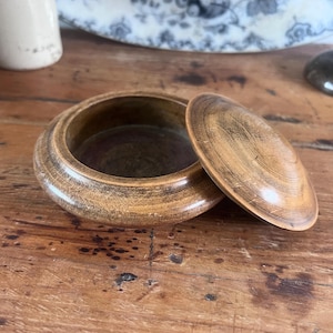 May include: A round, wooden trinket box with a lid, resting on a wooden surface. The box is open, revealing the interior. The wood has a warm, light brown color with visible grain patterns. The lid is slightly ajar, showing the inside of the box.