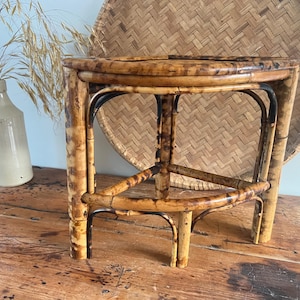 May include: A small, round bamboo side table with a dark, varnished finish. The table has three legs and a woven bamboo tray in the background. A ceramic vase with dried grasses is to the left. The table is on a wooden surface.