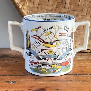 May include: A white ceramic mug with two handles, decorated with a colourful illustration of farming tools, people, and livestock. The mug has a blue rim and the words "GOD SPEED THE PLOUGH" on a banner.