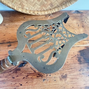 May include: A brass fireplace tool featuring an ornate, openwork lyre-shaped head with intricate floral and scroll detailing. The tool includes a cylindrical, ribbed wooden handle attached to the metal head, showcasing a metallic finish.