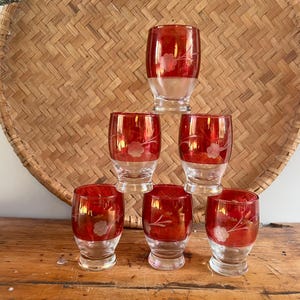 May include: Six vintage shot glasses with a red and clear glass design. Each glass has a floral etched pattern. The glasses are displayed on a wooden surface, in front of a woven basket.