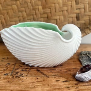 May include: White ceramic seashell-shaped bowl with a light green interior. The bowl has a textured, ridged exterior and a curved handle. The bowl is on a wooden surface with seashells and a woven background.