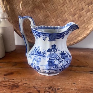 May include: A white and blue ceramic jug with a handle. The jug features a detailed blue landscape design. The handle and rim are also decorated with blue patterns. The jug is on a wooden surface.