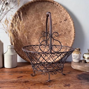 May include: A dark brown metal wire basket featuring a decorative scrollwork design, a single arched handle, and four small feet. The basket has an open lattice structure and a circular rim, providing a sturdy frame for holding various items.