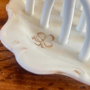 May include: A white ceramic utensil holder with a scalloped edge. The holder features a floral design in gold and has multiple vertical white bars for holding utensils. The holder is on a wooden surface.