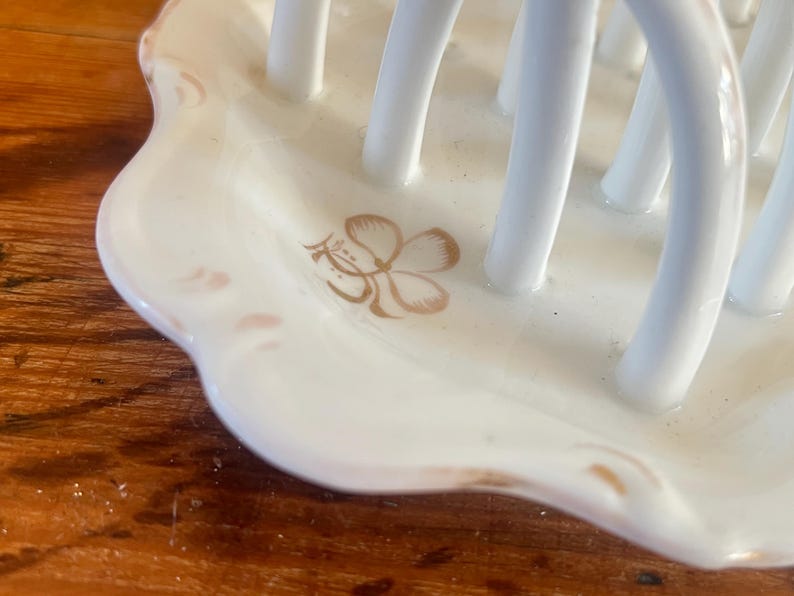 May include: A white ceramic utensil holder with a scalloped edge. The holder features a floral design in gold and has multiple vertical white bars for holding utensils. The holder is on a wooden surface.