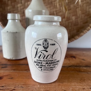 Peut inclure: Un pot en céramique de couleur crème avec un col cylindrique et un corps arrondi. L'avant présente un texte imprimé en noir indiquant : The Virol Ideal Food, Virol, A Preparation of Bone-Marrow, An Ideal Fat Food for Children & Invalids.