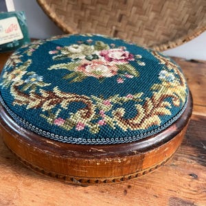May include: Circular footstool with a teal needlepoint top displaying a floral pattern in pink, white, and green. The wooden base is stained brown with decorative trim. A vintage piece for home decor.