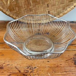 May include: A silver-toned wire fruit basket with a rounded base and a woven wooden insert. The basket has a unique, open design and sits on a wooden surface. A woven basket is in the background.