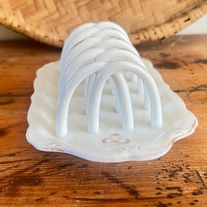 May include: White ceramic toast rack with arched dividers, set on a scalloped dish with a gold trim and a floral design. The rack is placed on a wooden surface, with a woven basket in the background.