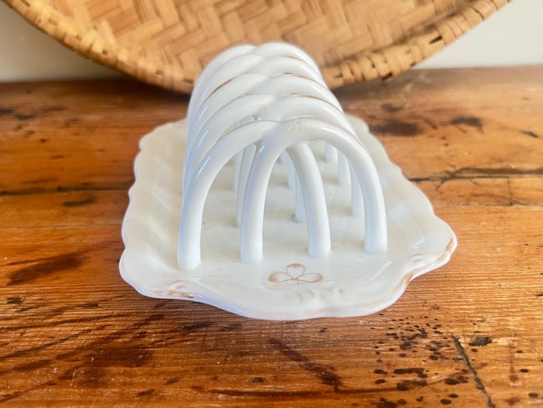 May include: White ceramic toast rack with arched dividers, set on a scalloped dish with a gold trim and a floral design. The rack is placed on a wooden surface, with a woven basket in the background.