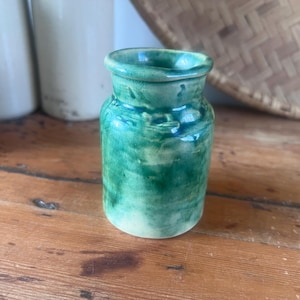 May include: A small ceramic jar with a glossy, mottled green glaze. The vessel features a cylindrical body with a slightly flared rim and a short, narrow neck. The surface displays variations in color intensity, ranging from deep forest green to pale lime.