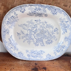 May include: An oval ceramic platter featuring a cream-colored base with a detailed blue floral transferware pattern. The design includes a central bouquet of various flowers and leaves, surrounded by a decorative border of floral motifs.