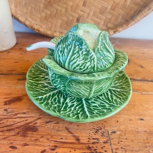 May include: A green ceramic cabbage-shaped serving dish with a lid and saucer. The dish features detailed leaf patterns. A small white spoon is on the lid. The set is designed to look like a cabbage, a charming kitchenware item.