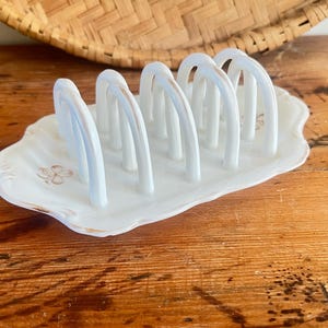 May include: A white ceramic toast rack with six arched slots. The rack has a scalloped edge and is decorated with a floral design. The base has a light brown trim. A woven basket is in the background.