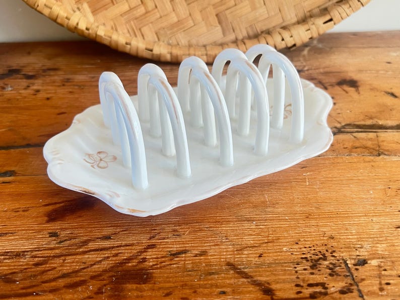 May include: A white ceramic toast rack with six arched slots. The rack has a scalloped edge and is decorated with a floral design. The base has a light brown trim. A woven basket is in the background.