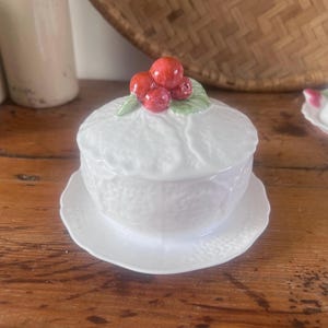 May include: White ceramic butter dish with a lid, on a matching saucer. The lid is topped with red cherries and green leaves. The dish has a textured, embossed design. A similar dish is in the background.