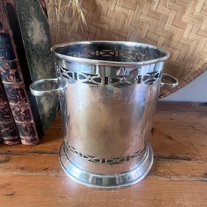 May include: A cylindrical silver-toned metal wine cooler or ice bucket featuring two side handles and decorative openwork bands with geometric patterns at the top and bottom rims. The surface has a polished finish with visible signs of patina.
