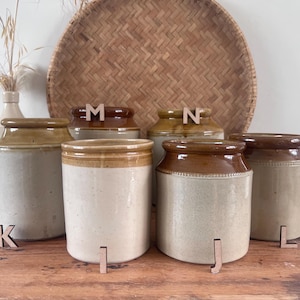 May include: A selection of antique ceramic jars in shades of brown and cream, displayed on a wooden table. The jars have a classic design, with wooden letter cutouts in front of some. A woven basket and a vase with dried flowers are in the background.