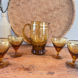 May include: Amber-colored glassware set, including a pitcher and four stemmed glasses, each with gold horizontal stripes. The set is arranged on a wooden surface, with a woven basket and a vase in the background.