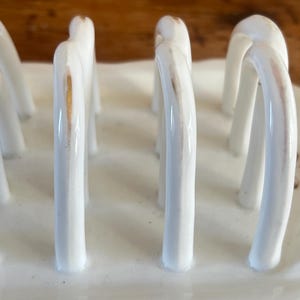 May include: A white ceramic toast rack with arched dividers. The rack has a rectangular base and features gold accents on the dividers. The design is simple and elegant, suitable for serving toast or bread.