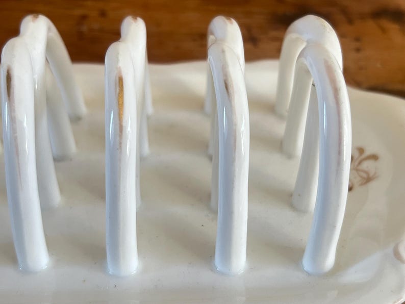 May include: A white ceramic toast rack with arched dividers. The rack has a rectangular base and features gold accents on the dividers. The design is simple and elegant, suitable for serving toast or bread.
