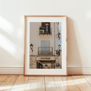 May include: Framed print of a building facade with balconies, shutters, and the text "BAR LA PLAZA". The building is painted in a light beige color, with a wooden frame. The image is set against a white wall and wooden floor.