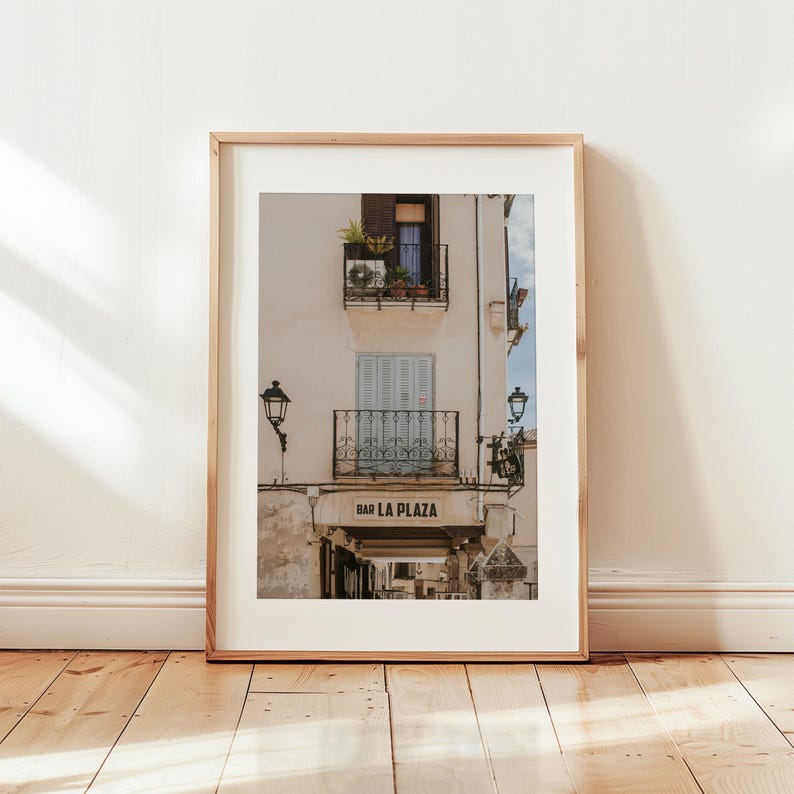 May include: Framed print of a building facade with balconies, shutters, and the text "BAR LA PLAZA". The building is painted in a light beige color, with a wooden frame. The image is set against a white wall and wooden floor.