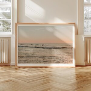May include: A framed photograph of a seascape with surfers riding waves at sunset. The water is a light blue and the sky is a soft pink and orange.