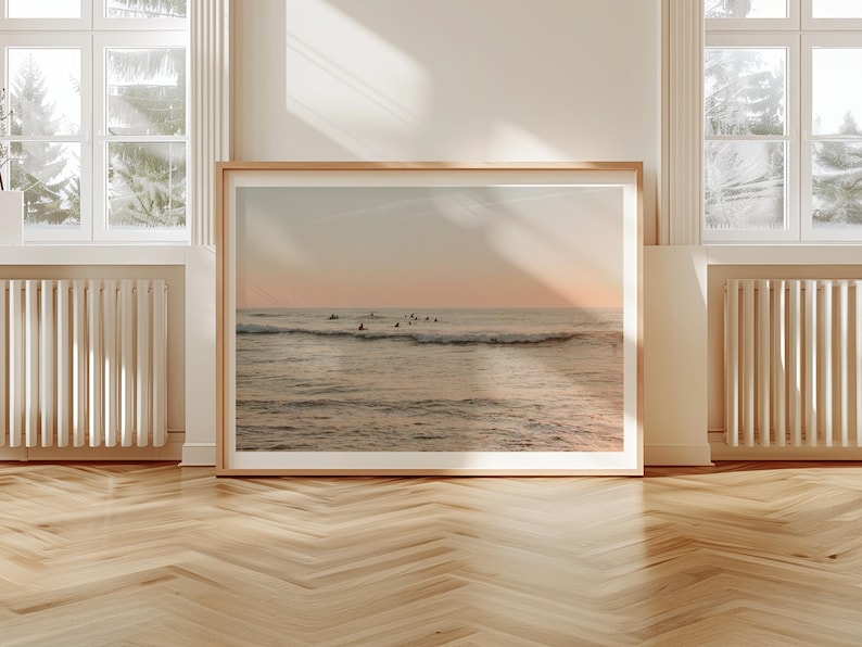 May include: A framed photograph of a seascape with surfers riding waves at sunset. The water is a light blue and the sky is a soft pink and orange.