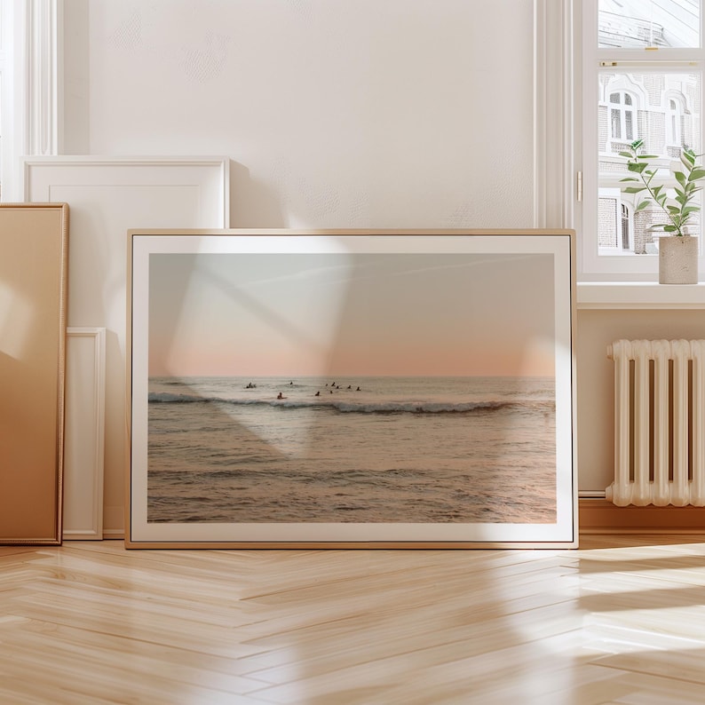 May include: A framed photograph of a seascape with a group of surfers paddling out in the ocean at sunset. The sky is a soft pink and orange, and the water is a calm blue.
