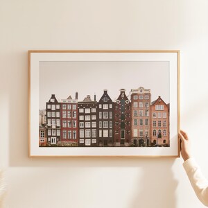 Puede incluir: Una impresi&oacute;n art&iacute;stica enmarcada que muestra una hilera de casas de canal estrechas de varios pisos con fachadas arquitect&oacute;nicas variadas en tonos rojo, marr&oacute;n y gris oscuro. Los edificios tienen m&uacute;ltiples ventanas con marcos blancos.