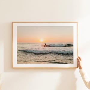 May include: A framed photograph of a surfer riding a wave at sunset. The sky is a vibrant orange and pink, and the ocean is a deep blue. The surfer is silhouetted against the setting sun.