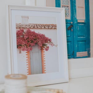 K&ouml;nnte beinhalten: Gerahmter Druck eines wei&szlig;en Geb&auml;udes mit einer hellblauen T&uuml;r und rosa Blumen, die dar&uuml;ber herabfallen. Der Rahmen ist wei&szlig;, und das Bild ist vor einem wei&szlig;en Hintergrund platziert. Ein zweites gerahmtes Bild einer blauen T&uuml;r ist sichtbar.