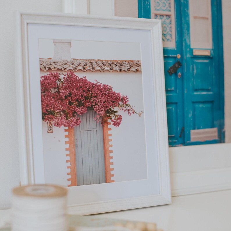 K&ouml;nnte beinhalten: Gerahmter Druck eines wei&szlig;en Geb&auml;udes mit einer hellblauen T&uuml;r und rosa Blumen, die dar&uuml;ber herabfallen. Der Rahmen ist wei&szlig;, und das Bild ist vor einem wei&szlig;en Hintergrund platziert. Ein zweites gerahmtes Bild einer blauen T&uuml;r ist sichtbar.