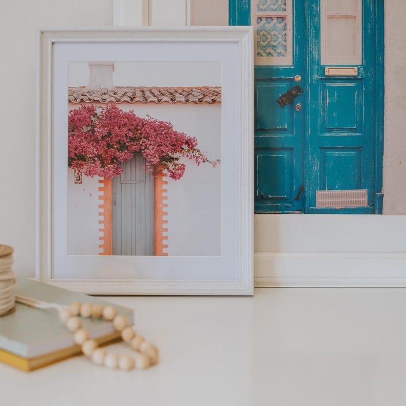 K&ouml;nnte beinhalten: Ein gerahmter Druck eines wei&szlig;en Geb&auml;udes mit einer grauen T&uuml;r und leuchtend rosa Blumen. Der Rahmen ist wei&szlig;, und das Bild ist vor einem wei&szlig;en Hintergrund platziert. Ein zweites Bild einer blauen T&uuml;r ist sichtbar.