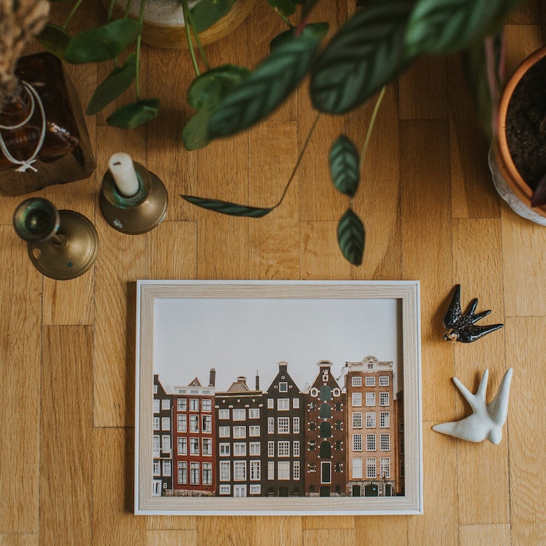 Puede incluir: Un marco de fotos rectangular que contiene una impresi&oacute;n de una hilera de casas de canal estrechas y de varios pisos con fachadas a dos aguas variadas en tonos rojo, marr&oacute;n y bronceado. El marco es de madera clara con un perfil simple.