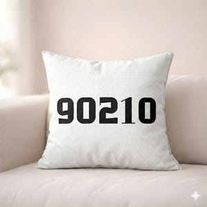 Puede incluir: Cojín cuadrado blanco con el número "90210" en negro impreso en la parte delantera. El cojín está sobre un sofá rosa claro. Los números están en negrita, fuente sans-serif.