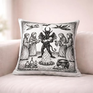 May include: Square decorative pillow with a black and white illustration depicting a ritual scene. A horned figure holds a baby over a fire, surrounded by robed figures, bottles, and a cauldron. The pillow is on a pink surface.