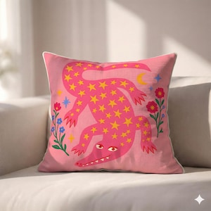 May include: A square throw pillow with a pink fabric cover featuring a central illustration of a pink alligator covered in yellow stars. The design includes small red and blue flowers on the sides, a yellow crescent moon, and blue stars.