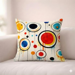 May include: Square throw pillow with a colorful abstract design. The pillow features a cream-colored background with a pattern of overlapping circles in various sizes and colors, including red, blue, yellow, and black. The circles are outlined in black and have a watercolor effect.