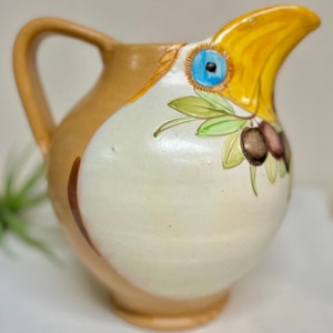 May include: A ceramic pitcher with a bird-shaped spout and a hand-painted design of olives and leaves. The pitcher is brown with a white glaze and yellow, green, and blue accents.