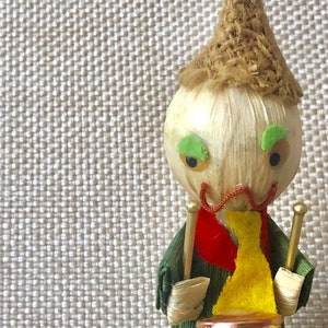 May include: A handmade corn husk doll wearing a green dress, a yellow scarf, and a brown hat. The doll is holding a small drum and drumsticks.